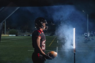 American football player is holding a ball, wearing a helmet and uniform, and standing on a playing