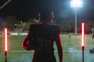 Football player stands on stadium field at night, helmet and uniform on, holding ball and facing