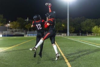American football players wearing helmets and jerseys leaping to catch a football during a night