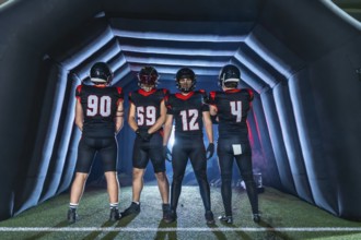 American football team in black uniforms stands inside an inflatable tunnel, helmets on, focused
