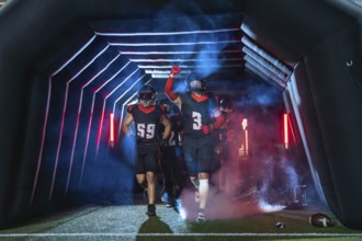 American football players burst from a stadium tunnel into smoke and colored lights, fists raised,