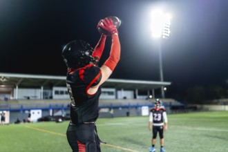American football player in red and black uniform and helmet stretches arms to catch ball under