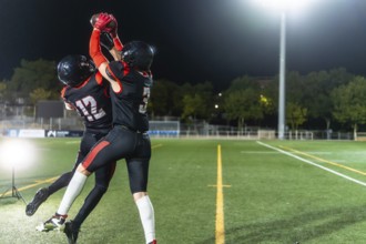 Two female american football players in black uniforms leap to catch the ball during a night game