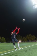 American football player in uniform and helmet is jumping to catch a football during a night game
