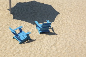 Teal blue wooden Adirondack chairs and casted shadow from parasol in fine tan sand, Clock Tower