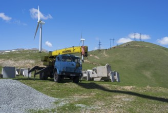 A blue crane lorry stands on a green construction site next to a wind turbine on a hill, Pushkin