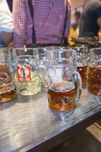 Large glass mugs of beer on a wooden table surface in a lively festival setting, Cannstatter Wasen