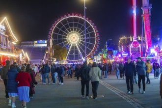 People walking on a festive square with a glowing Ferris wheel, Cannstatter Wasen folk festival,