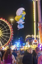 View of a Smurf balloon floating between a crowd at a funfair, with Ferris wheel in the background,