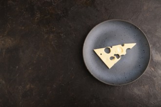 Piece of cheese with holes on blue plate on black concrete background, top view, flat lay, copy
