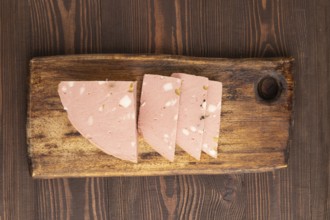 Mortadella sausage on wooden cutting board with pepper and herbs on brown wooden background. Top