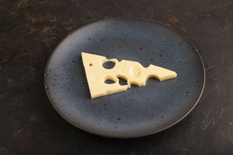 Piece of cheese with holes on blue plate on black concrete background, side view, close up,