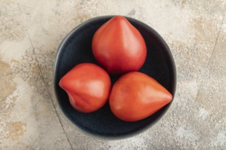 Red Heart shape tomatoes in blue bowl on brown concrete background. Top view, copy space, flat lay.