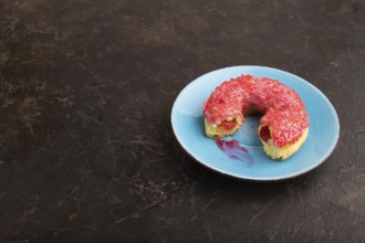 Bitten Pink Donut with sprinkles on blue ceramic plate on black concrete background, side view,