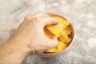 Dried Mango in ceramic bowl with hand on brown concrete background. Side view, close up. healthy