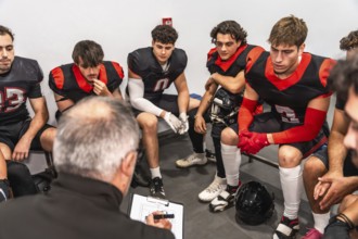 Football coach reviews clipboard plays while focused young players sit in the locker room,