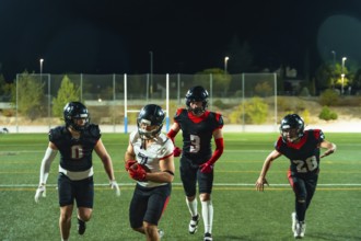 Four young american football players are running on the field under stadium lights at night, with