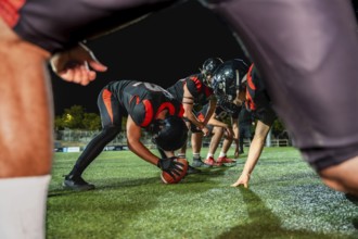 American football players line up on the field at night, a center snapping the ball, preparing for