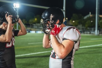 American football players adjusting their helmets on illuminated green field, preparing for intense
