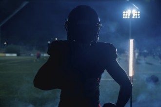 American football player standing in silhouette on a dark field under bright stadium lights at