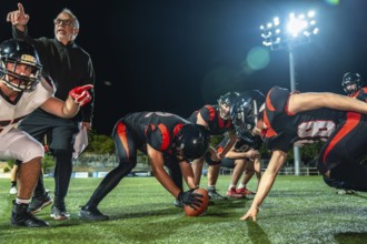 American football team huddled on a dark stadium field under bright lights at night, coach pointing