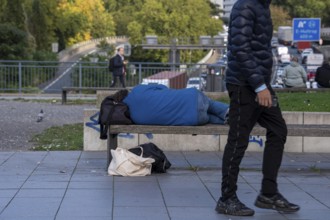 Man sleeping on a bench, travelling bag serves as pillow, in the city centre, passers-by walk by