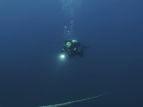Ein Taucher mit Lampe steigt aus der dunklen Unterwasserwelt auf, einem Seil folgend. Tauchplatz