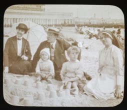 Affluent Edwardian family sitting on sandy seaside beach man, woman, children with nursemaid