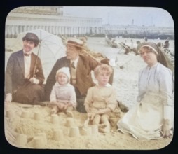 Affluent Edwardian family sitting on sandy seaside beach man, woman, children with nursemaid
