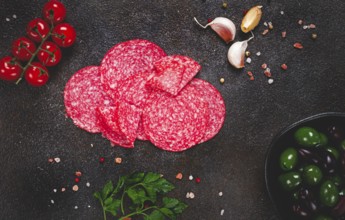 Composition of sliced salami, with fresh tomatoes, garlic and green olives, on a dark surface,