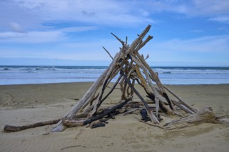 Treibholzstruktur in Zeltform am Strand unter blauem Himmel und Meerblick, Kaka Point, Clutha