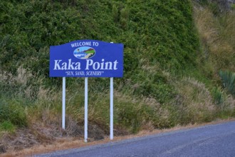 Willkommensschild von Kaka Point neben einer Straße mit grüner Vegetation und landschaftlicher