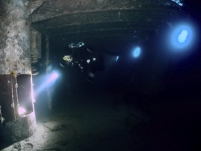 Diver exploring a dark wreck underwater, illuminated by torchlight. Dive site wreck of the Vis,