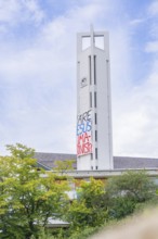 Church tower with protest banner between trees under blue sky, Tesla Model Y Juniper, Gechingen,