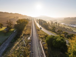 Landscape at sunrise with parallel railway tracks and country road, surrounded by fields and trees,