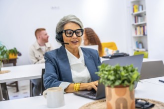 Senior woman actively working in a modern office, wearing a headset and spectacles while typing on