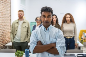 Young man standing with arms crossed, confidently looking at the camera, with his diverse team
