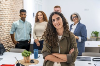 Confident young woman with curly hair smiling at camera, standing with arms crossed in a modern