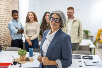 Experienced senior woman standing confidently in blue blazer and glasses, leading and collaborating