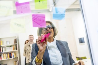 Mature businesswoman attaching colorful sticky notes on a clear glass board, collaborating on