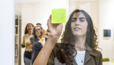 Young businesswoman sticking a green sticky note on a transparent glass wall during a team meeting,