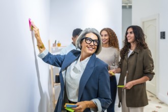 Confident gray haired businesswoman in glasses posts a sticky note during a lively team