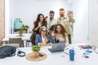 Diverse business colleagues smiling and waving at a laptop during a video call, connecting with