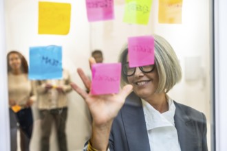 Senior business woman smiling, organizing colorful sticky notes on a glass whiteboard during a