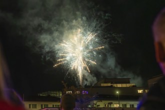 Colourful fireworks light up the night sky over a dark building complex with spectators, Fire