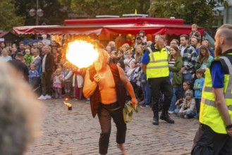 A fire eater entertains a crowd on a cobbled street in front of red stalls, Fire Night,