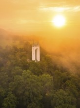 White tower rises out of misty forest while the sun illuminates orange sky, Schönbergturm,