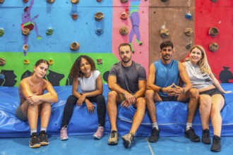 Five athletes resting on a vibrant blue crash pad in a colorful bouldering gym, enjoying a well