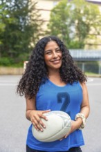 Confident female rugby player is smiling while holding a rugby ball, standing on a sports field,