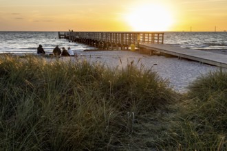 People sitting on the beach while a long pier leads into the sea, framed by sunset lights, Kobaek